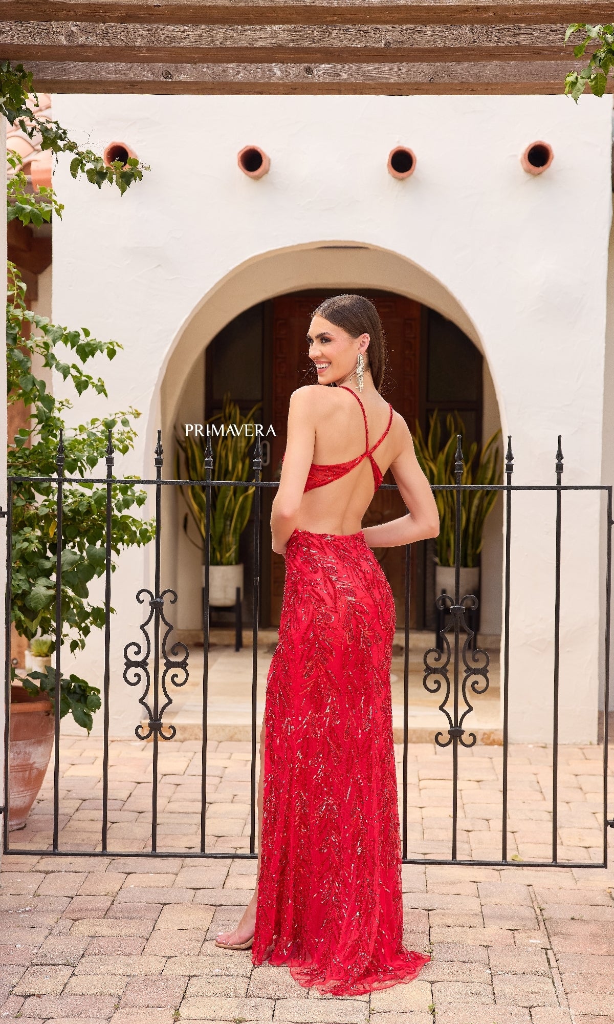 A woman stands outdoors by a gate, smiling over her shoulder in the Primavera 4316 Open-Sides Long Sequin Prom Dress, featuring thin straps and a chic strappy-back design.